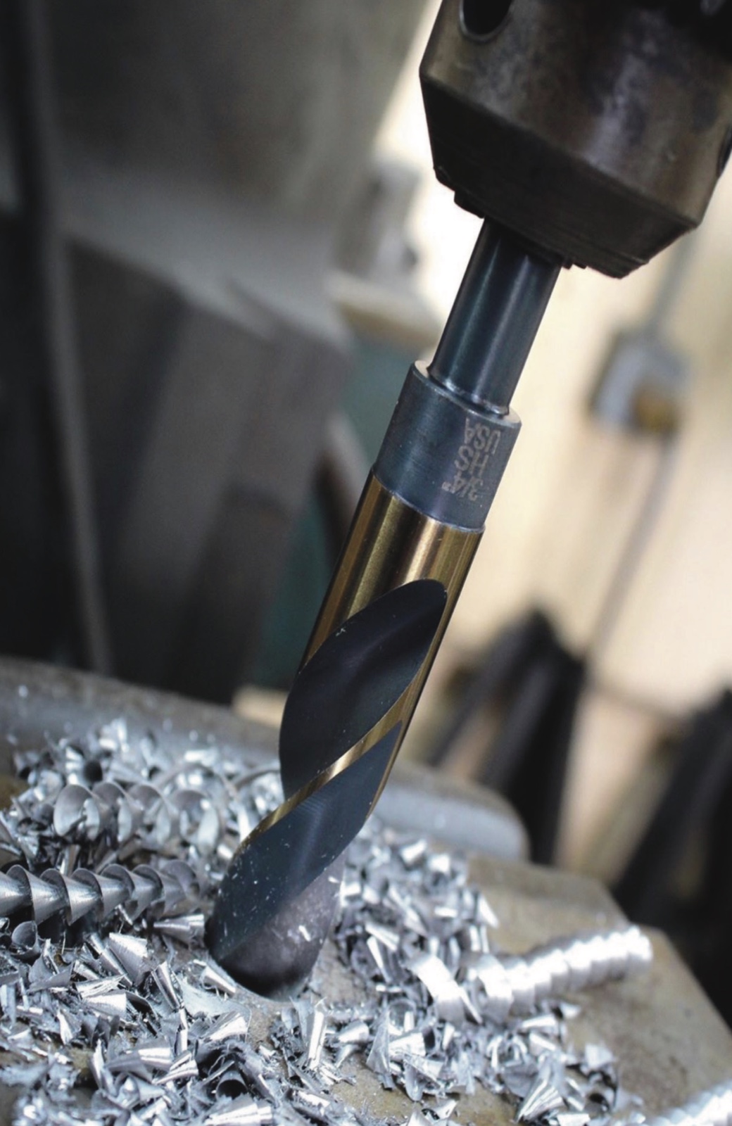 Black and gold 3/4 in. Silver and Deming drill bit cutting steel in a drill press, curled chips climbing out along the flutes