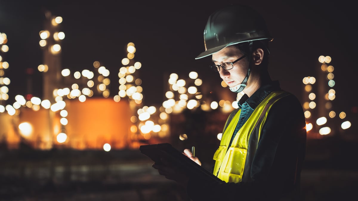 Civil engineer in a hard hat and safety vest reviewing plans on a tablet at night, plant lights behind