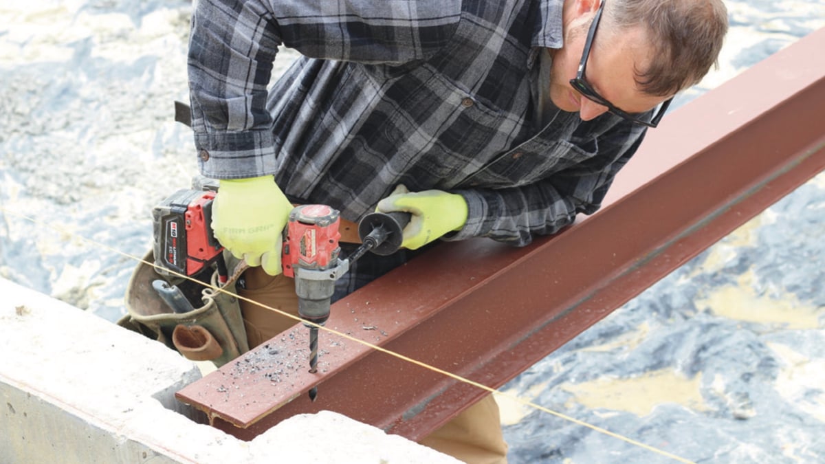 Worker drilling a steel I-beam flange with a cordless drill, metal chips on the beam