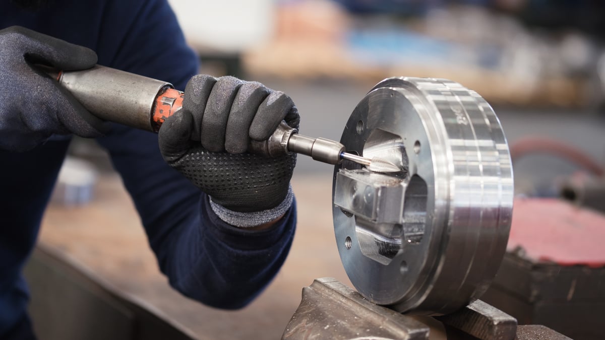 Machinist running a carbide burr in a die grinder on a bright metal part held in a vise